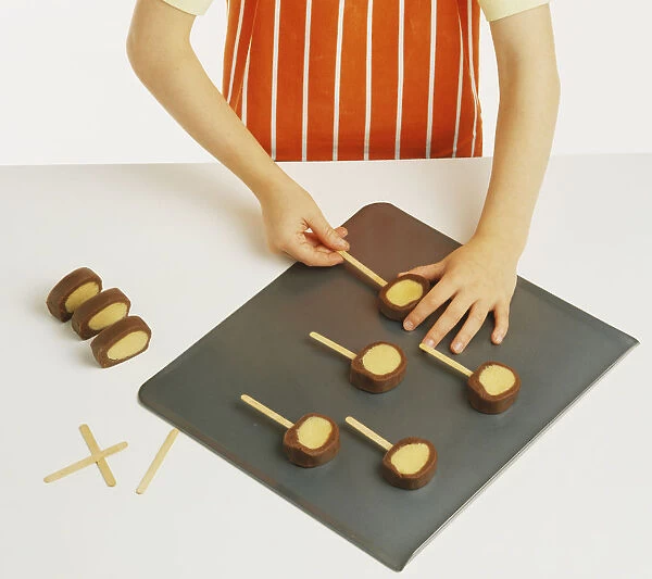 Hand model wearing orange and white striped apron, pushing lollipop sticks into chocolate