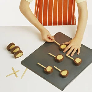 Hand model wearing orange and white striped apron, pushing lollipop sticks into chocolate