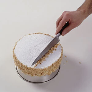 Man using kitchen knife to arrange chopped hazelnuts on top meringue gateau on baking tin